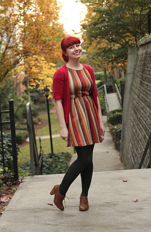 Woman in a fall-colored chevron dress with a short-sleeve red cardigan, dark tights and oxford heels