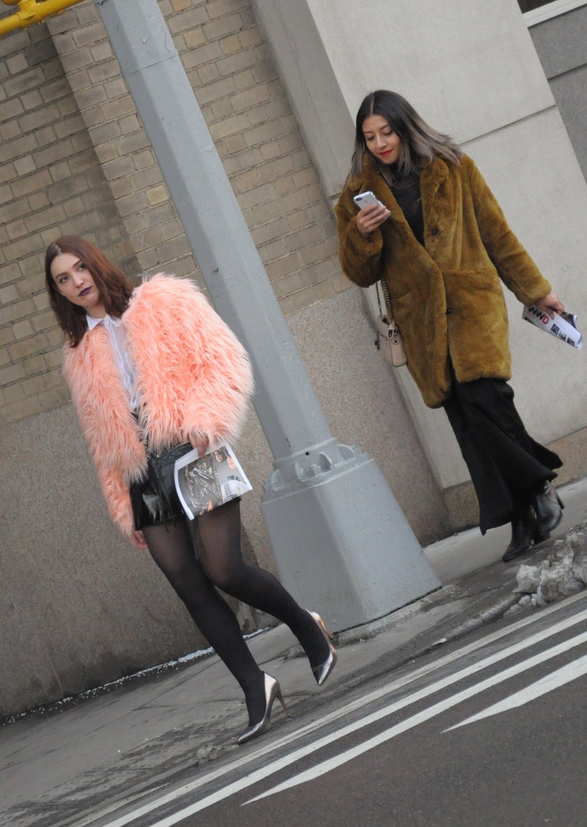 Two women walking on a city street wearing shaggy faux fur coats in pink and mustard over black outfits