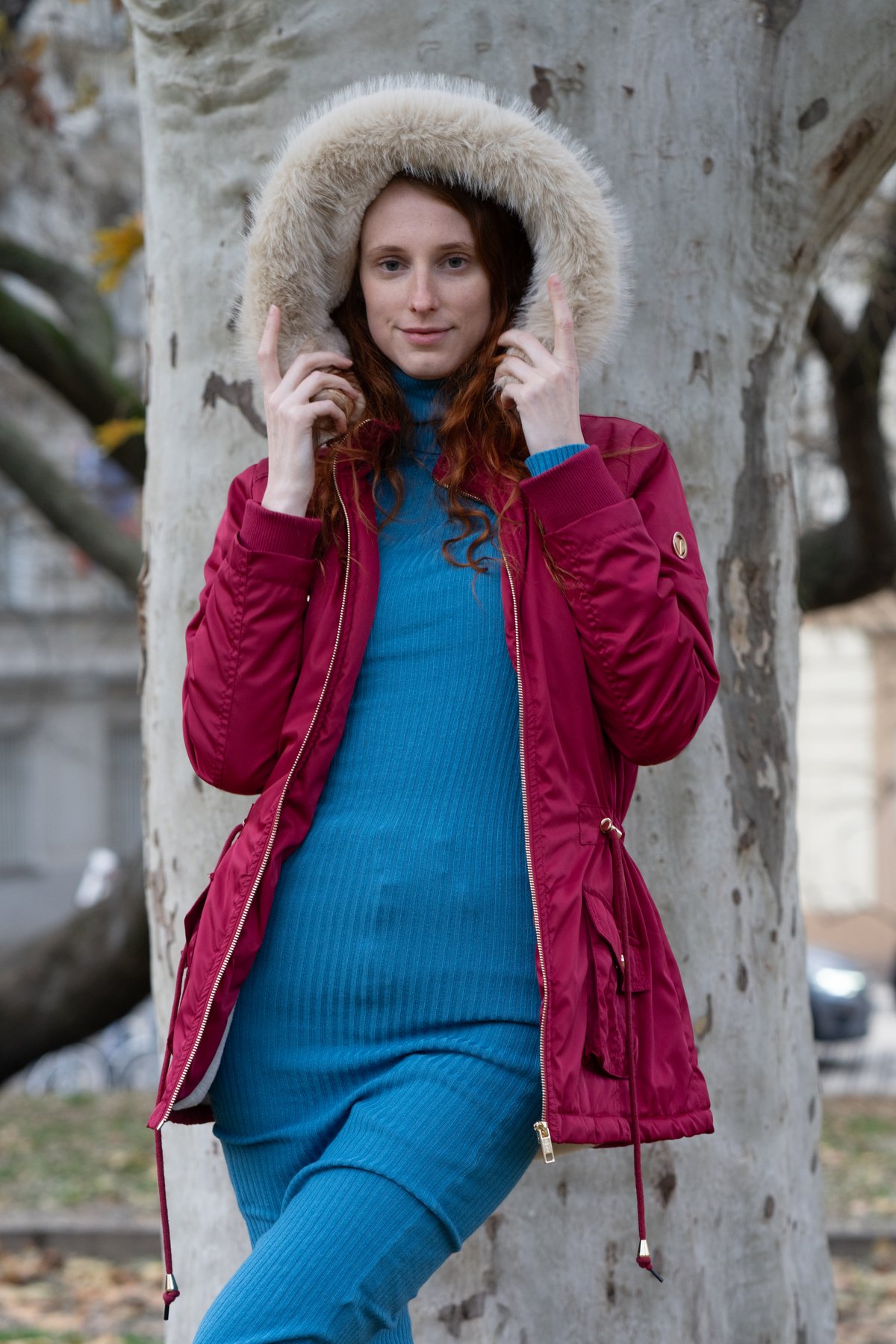 Young woman with long red hair holding up the cream faux-fur hood of a burgundy quilted coat worn open over a blue ribbed knit sweater dress