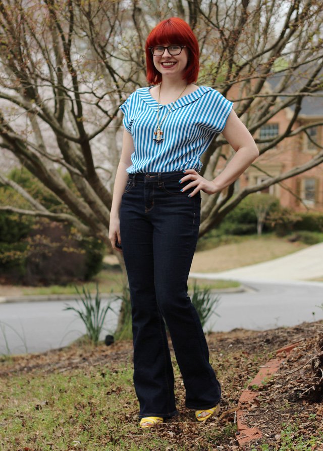 A woman with a red pixie haircut wearing a blue and white striped short-sleeve shirt, dark high-waisted flare jeans and yellow wedge sandals, standing outdoors by a tree
