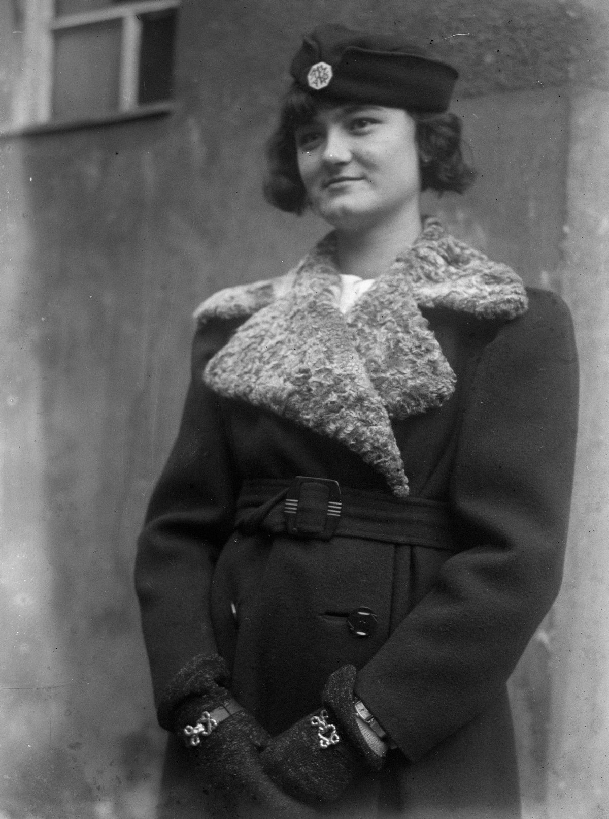 A three-quarter portrait of a woman in a dark belted coat with a large curly-lamb fur collar, a small dark cap with a brooch and embroidered dark gloves.