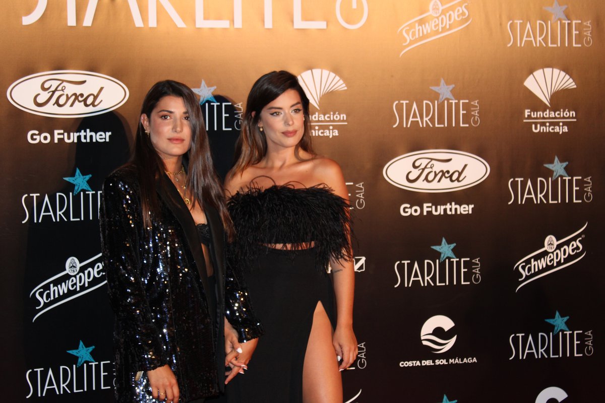 Two women at a gala in black sequin evening dresses, one strapless with a thigh slit