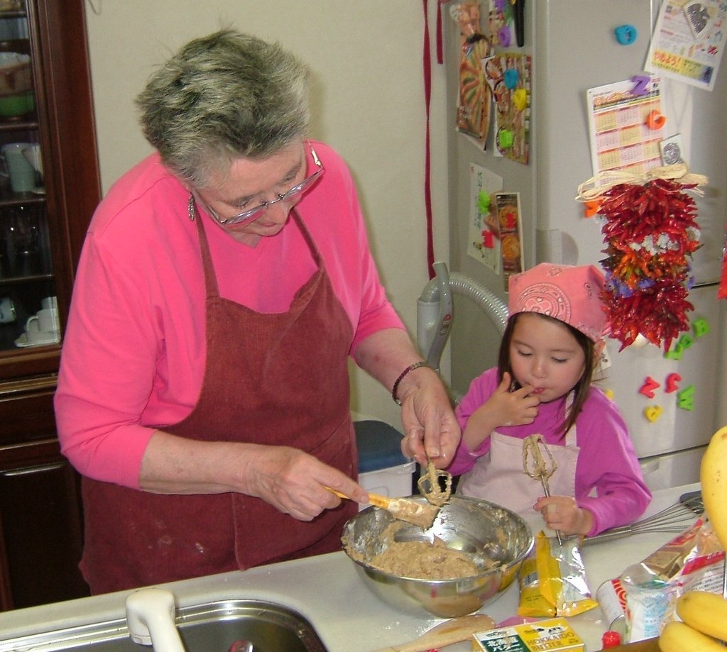 An older woman with glasses wearing a pink top and a red apron, mixing cookie dough in a metal bowl beside a young girl in a pink shirt in a home kitchen