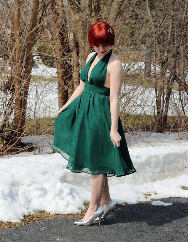 A woman with red hair wearing an emerald green chiffon halter dress and silver heels, standing in snow