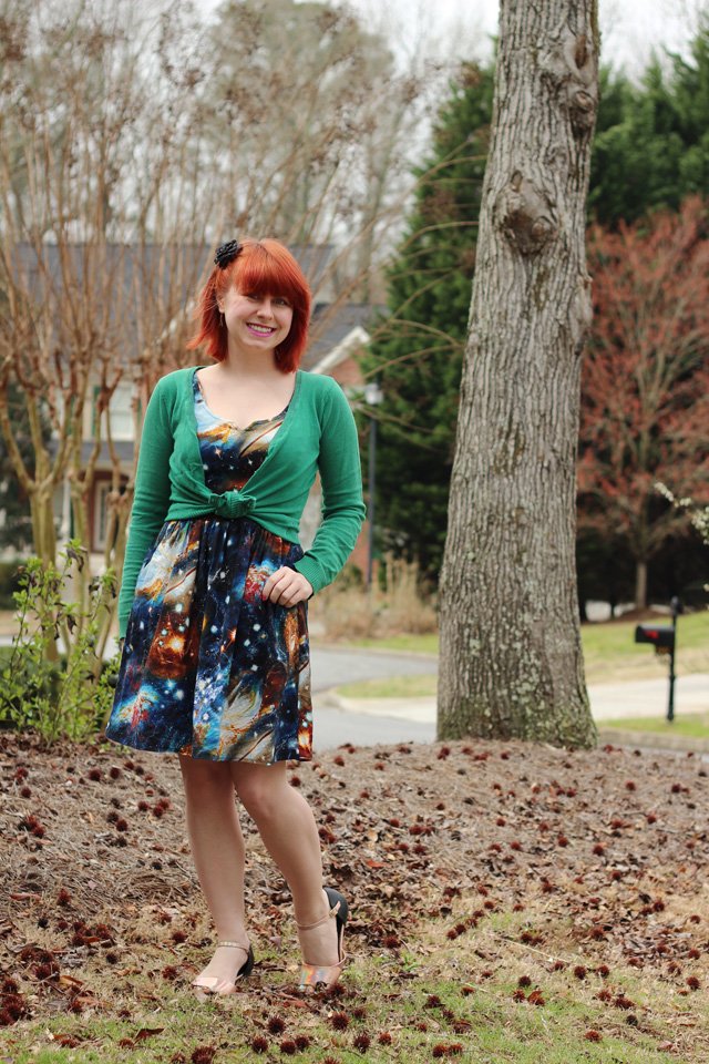 A smiling red-haired woman in a green knit cardigan tied at the front over a galaxy-print fit-and-flare dress and strappy heels, standing among trees