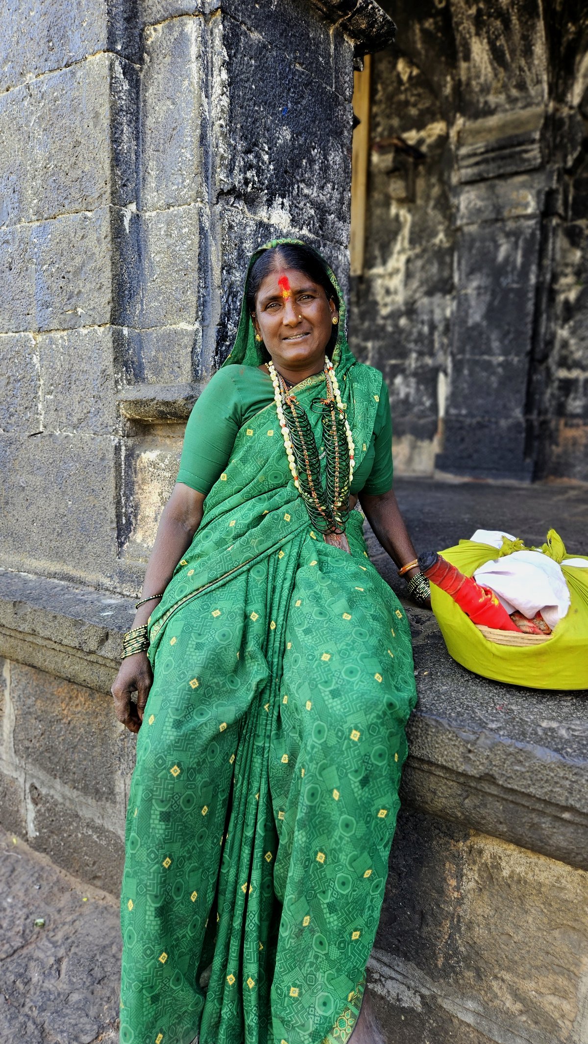 Woman seated outside a stone temple wearing a green printed cotton saree with green glass bangles and layered necklaces