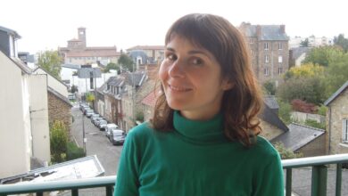 Woman with shoulder-length brown hair standing on a balcony overlooking a European town street, wearing an emerald-green turtleneck sweater with a clearly visible high folded neckline.