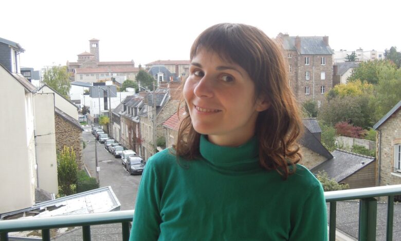 Woman with shoulder-length brown hair standing on a balcony overlooking a European town street, wearing an emerald-green turtleneck sweater with a clearly visible high folded neckline.