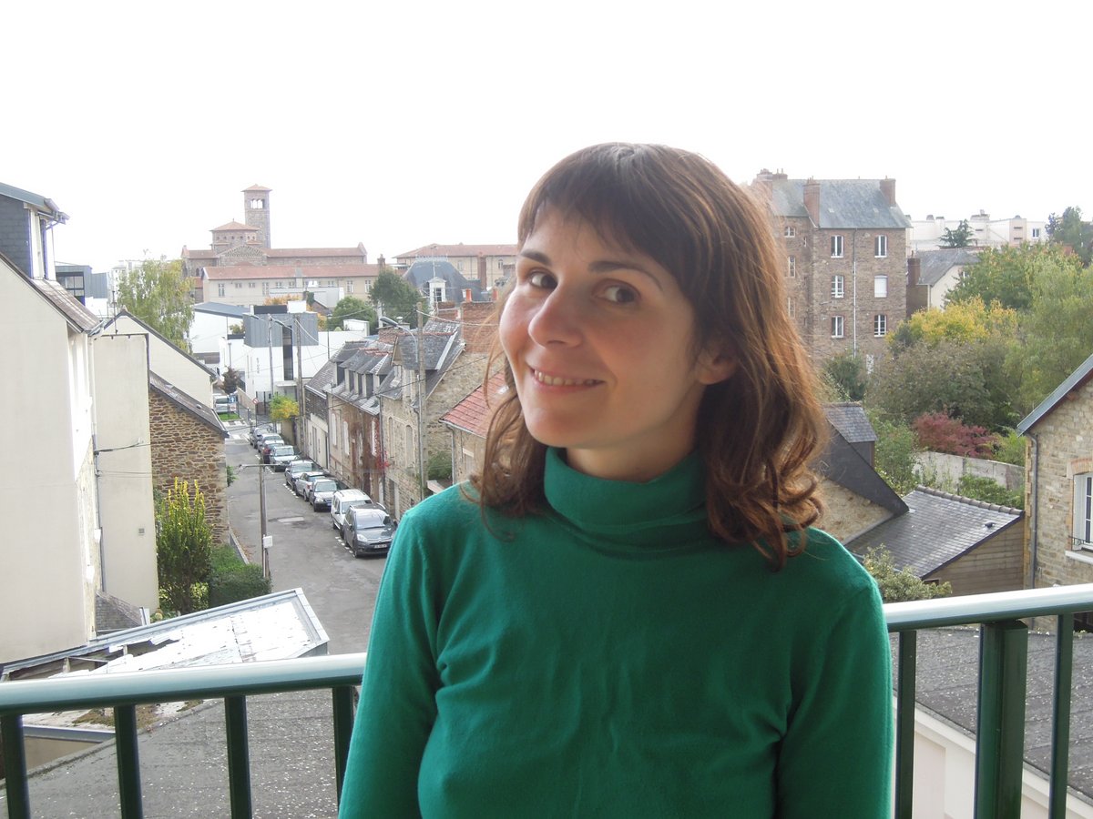 Woman with shoulder-length brown hair standing on a balcony overlooking a European town street, wearing an emerald-green turtleneck sweater with a clearly visible high folded neckline.