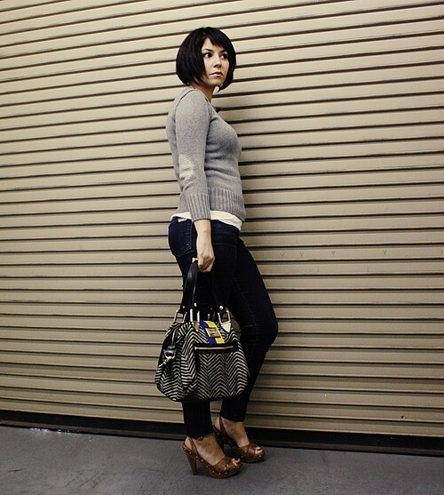 Woman in a grey knit sweater with dark skinny jeans, heels and a patterned handbag