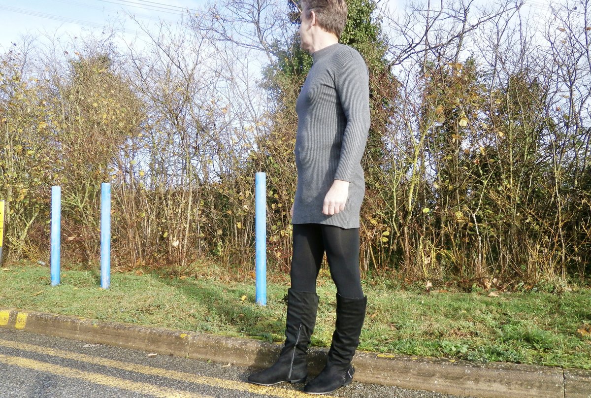 A model wearing a grey ribbed knit mini sweater dress with black opaque tights and calf-length black suede boots, photographed full-length against a plain background.
