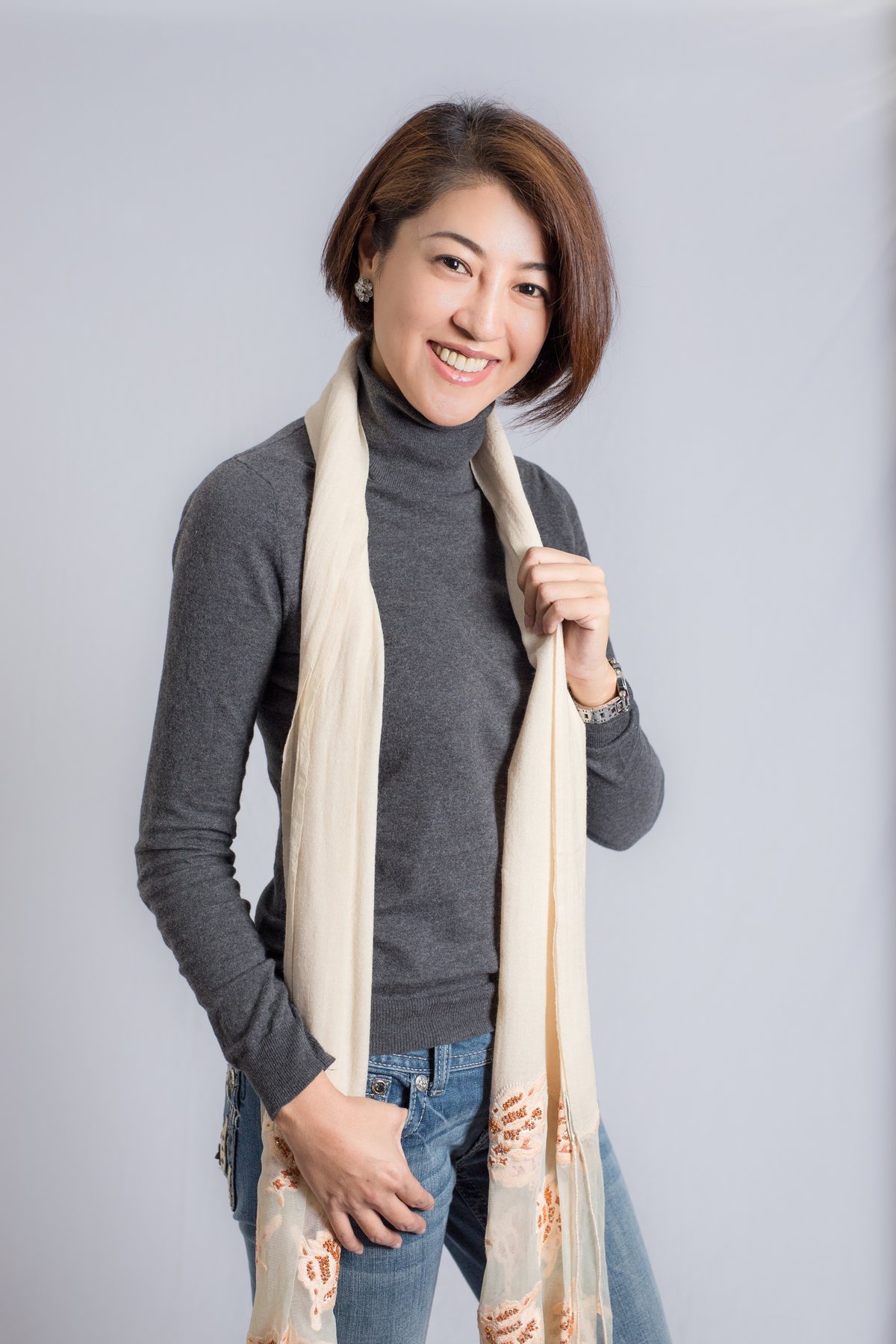 A studio portrait of a smiling woman wearing a grey turtleneck sweater, a long cream patterned scarf and blue jeans