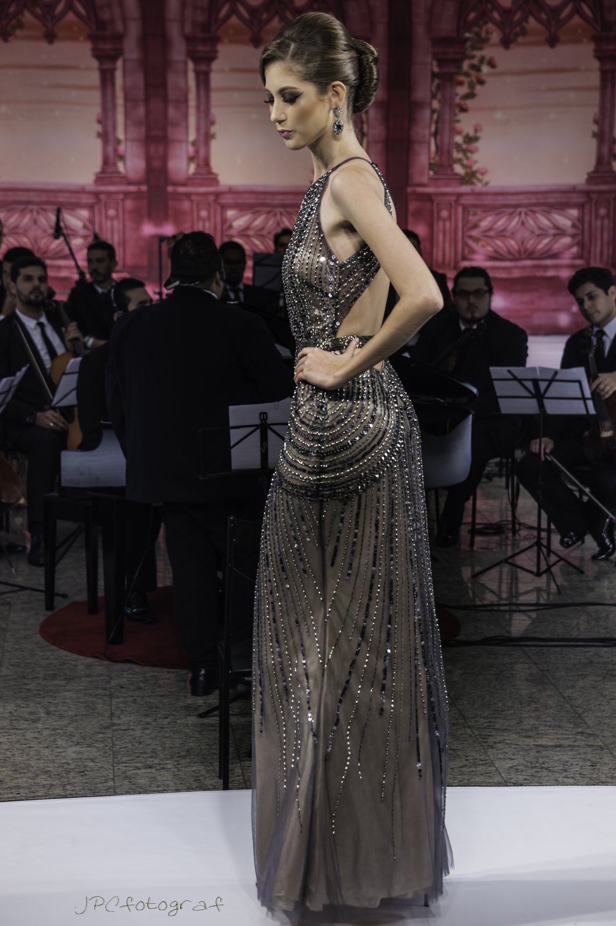 Model on a runway wearing a gunmetal sequin evening gown with dangling sequin strands and an open back