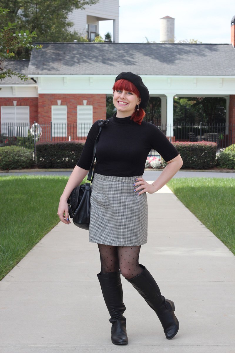 A red-haired woman on a path wearing a black beret, a black turtleneck top, a grey houndstooth mini skirt, dotted black tights and black knee-high boots with a black shoulder bag