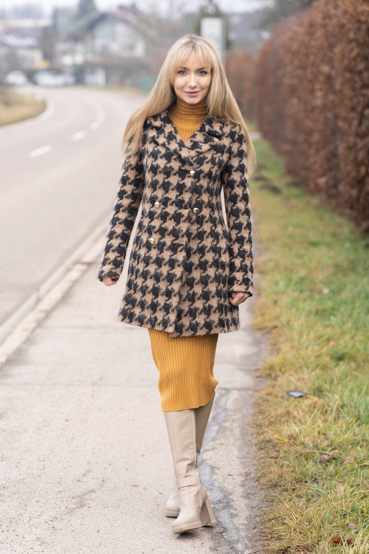 Smiling blonde woman walking outdoors wearing a tan-and-black houndstooth wool coat over a mustard ribbed knit dress with cream knee-high boots