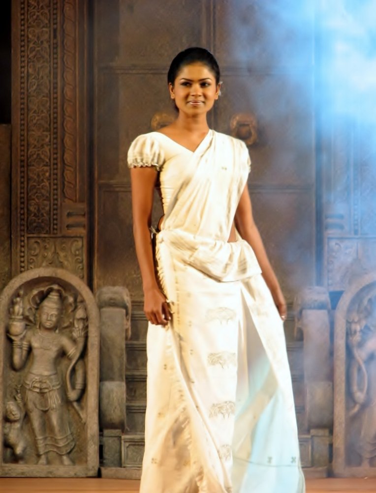 A young woman standing full-length on a stage wearing an ivory-white traditional Kandyan saree with subtle woven motifs and a puff-sleeved blouse, carved stone panels behind her