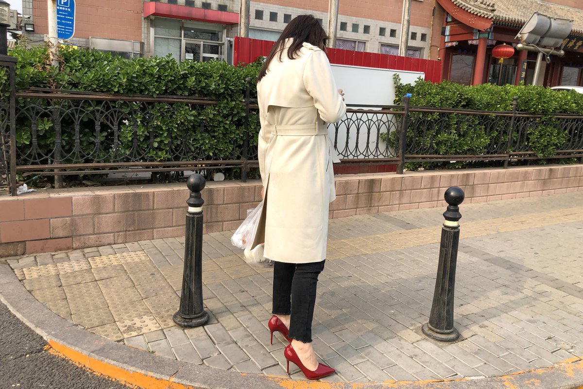 A woman photographed from behind on a Beijing street wearing an ivory belted trench coat, cropped dark jeans and glossy red patent pumps