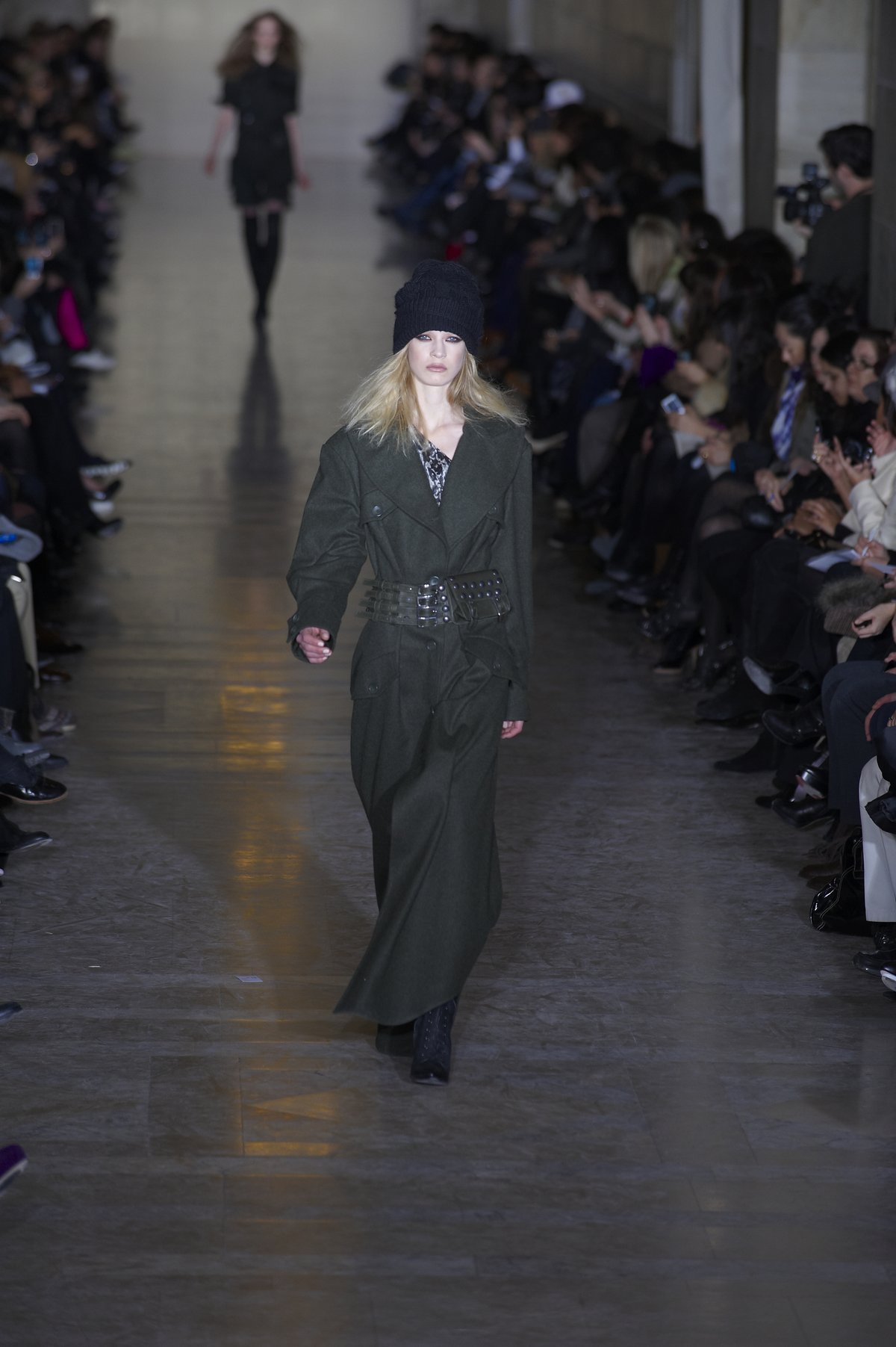 A model on a runway wearing a long dark belted wool coat with a wide studded belt, a knit beanie and a patterned scarf