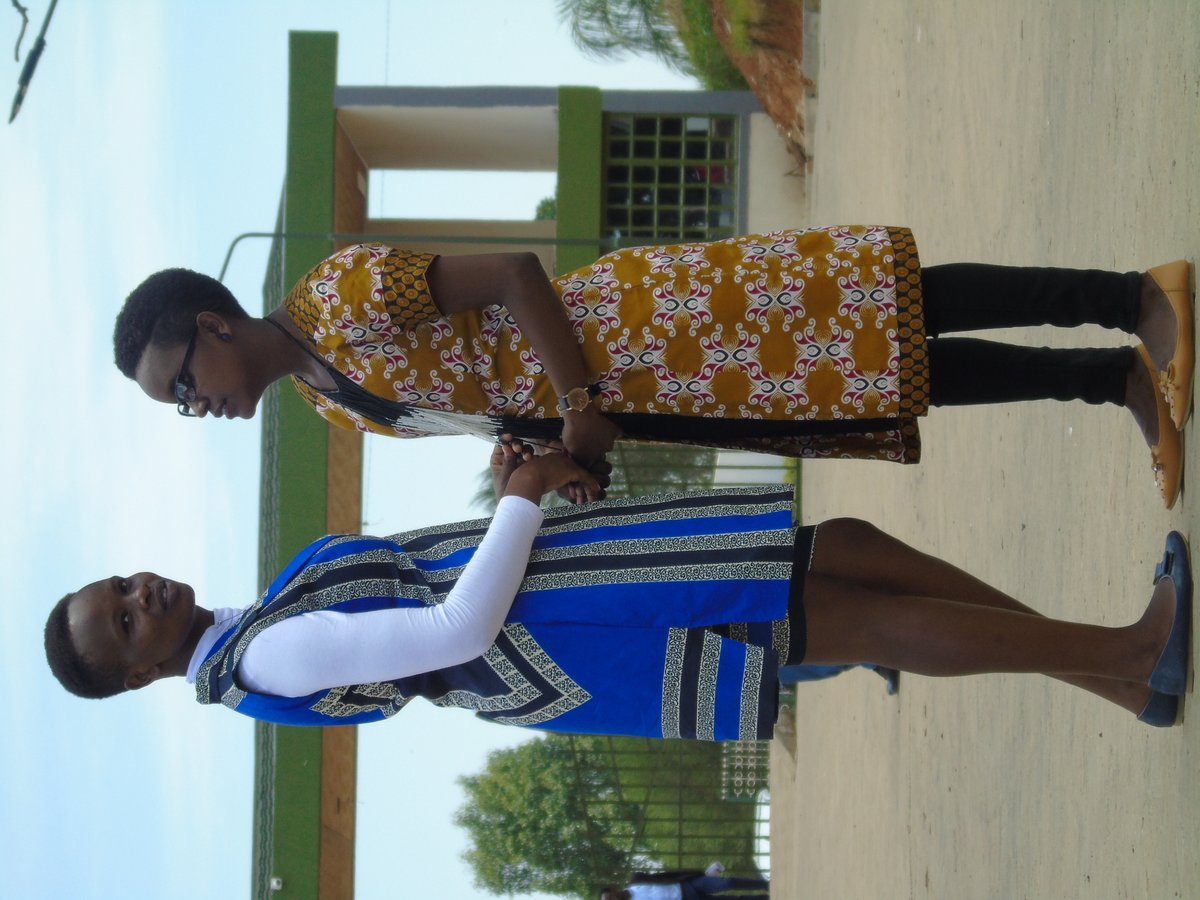 Two women standing outdoors wearing trench-style coats in blue and mustard African wax-print fabrics, one in stripes and one in an ornate floral pattern