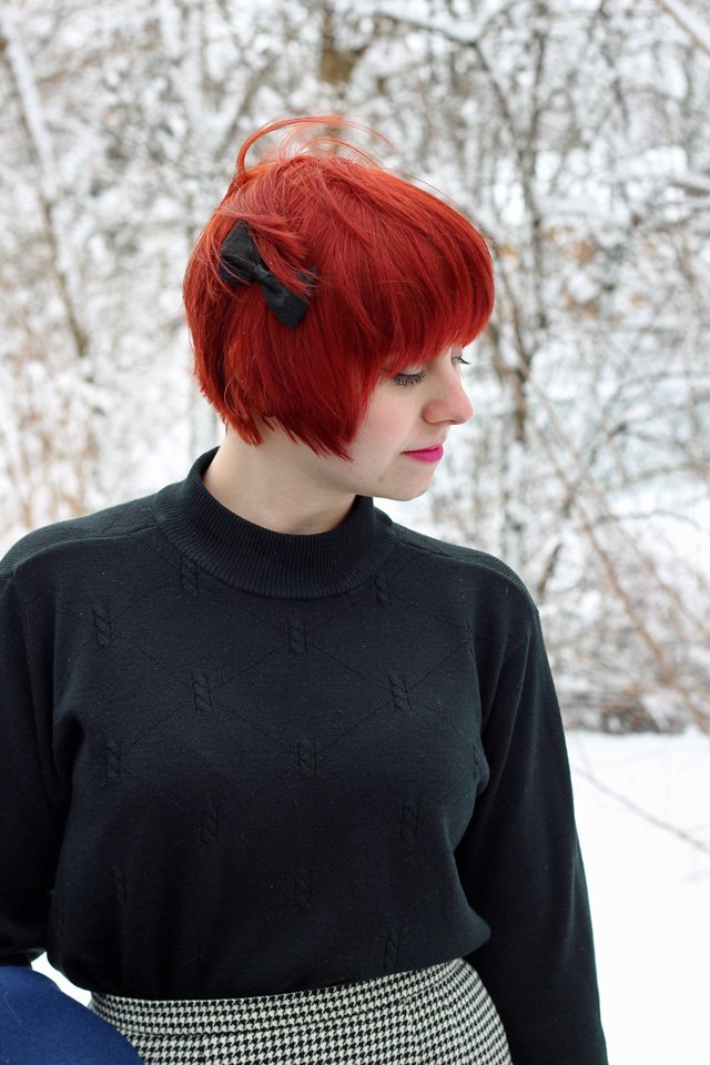 Close-up of a young woman with a short red pixie haircut and a black lace bow in her hair, wearing a black mock-neck textured knit sweater with the high collar clearly visible, snowy trees in the background.