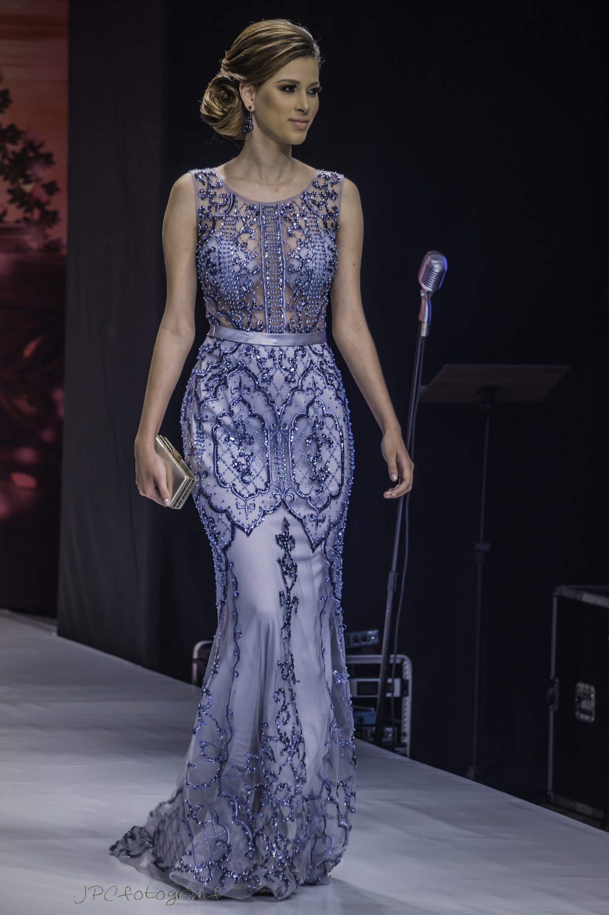 Model on a runway wearing a lavender-blue mermaid evening gown with blue beadwork and a satin waistband, holding a metallic clutch