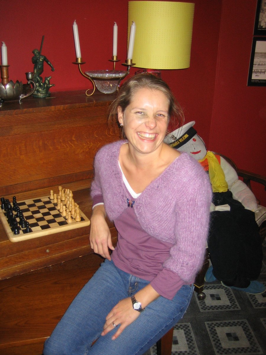 A smiling woman seated indoors wearing a cropped lilac-mohair bolero shrug over a purple v-neck top with blue jeans