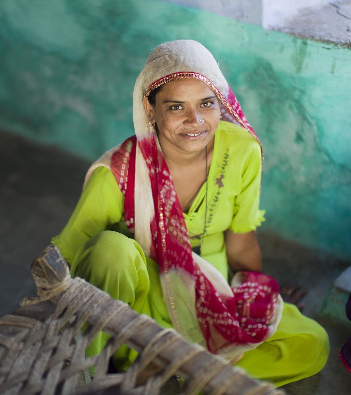 Woman seated on a woven cot wearing a lime green kurta and salwar with a red and white bandhani dupatta over her head