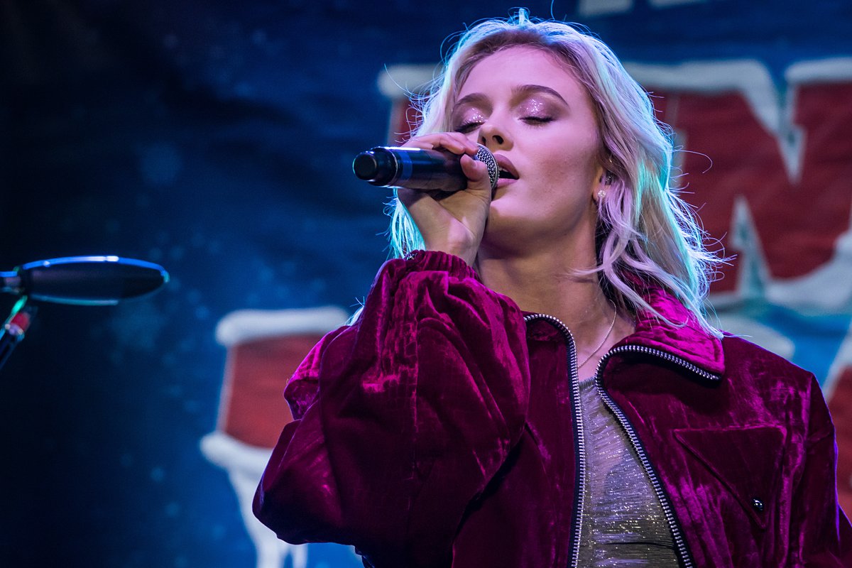 Zara Larsson performing on stage wearing a magenta crushed-velvet zip bomber jacket over a shimmery top, singing into a microphone.