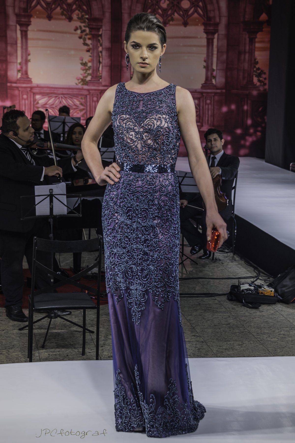 Model on a runway wearing a midnight blue beaded evening gown with silver scrollwork and an open back, holding a red clutch