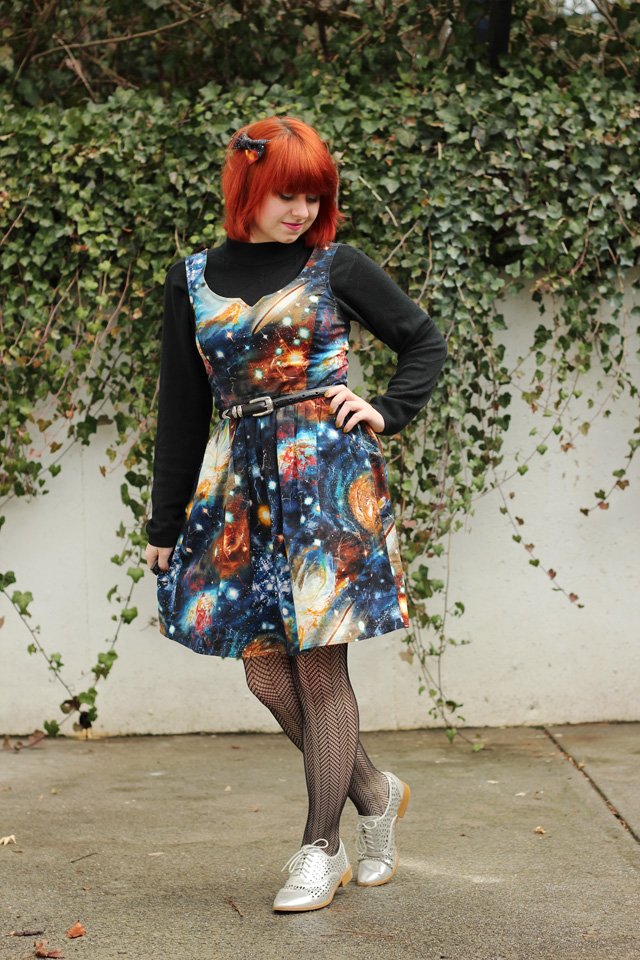 Full-length photo of a smiling young woman with short red hair wearing a black mock-neck top layered under a sleeveless galaxy-print fit-and-flare dress with a thin belt, black fishnet tights, and silver lace-up brogues, standing in front of an ivy-covered wall.