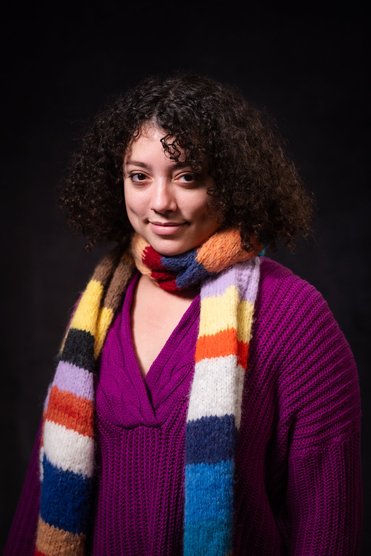 A studio portrait of a curly-haired woman wearing a magenta cable-knit sweater with a bold multicolored striped chunky knit scarf wrapped at her neck