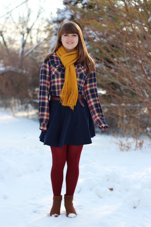 Young woman in snow wearing a mustard-yellow fringed scarf with a plaid shirt, navy skirt and burgundy tights