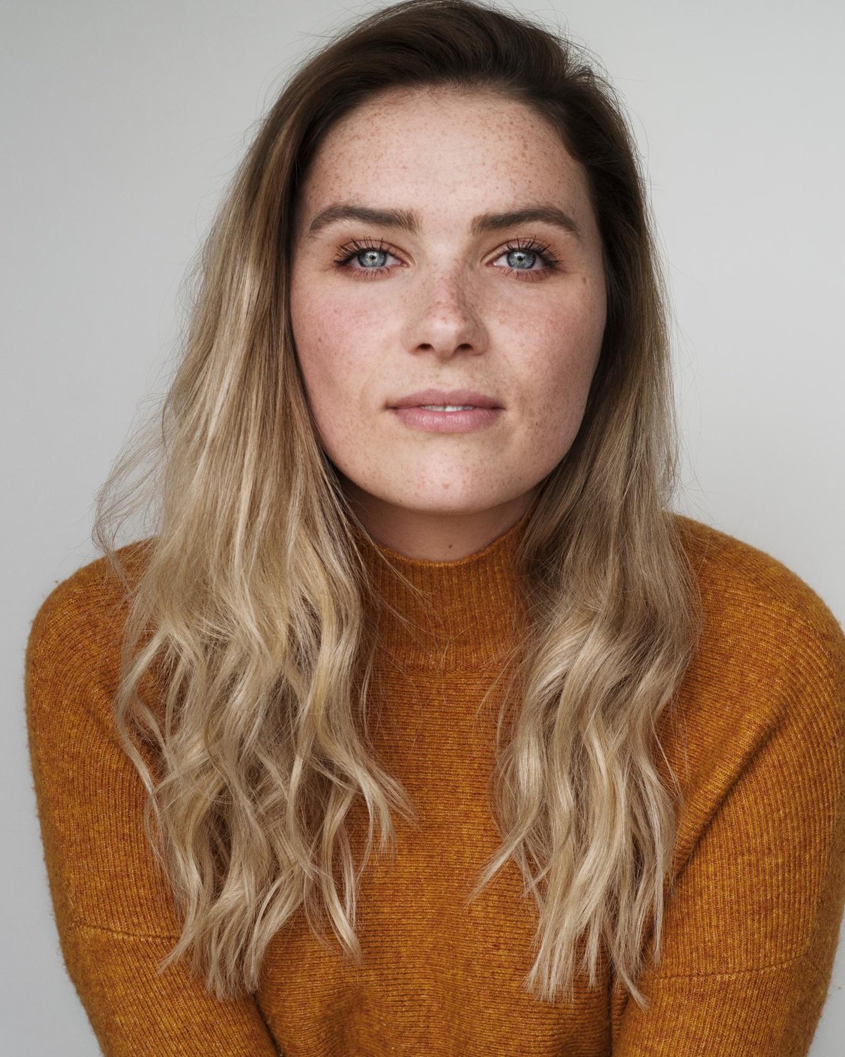 Close portrait of a blonde woman wearing a mustard-rust ribbed knit turtleneck against a light background.