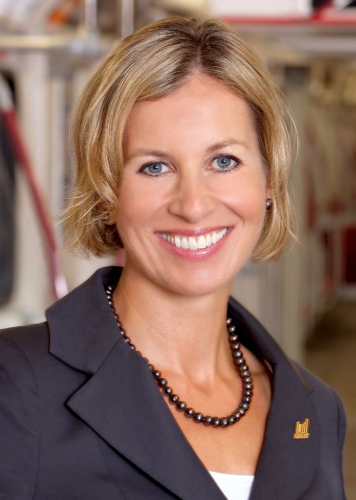 A smiling blonde woman in a professional headshot wearing a dark navy blazer over a white top with a dark pearl necklace and a small gold pin
