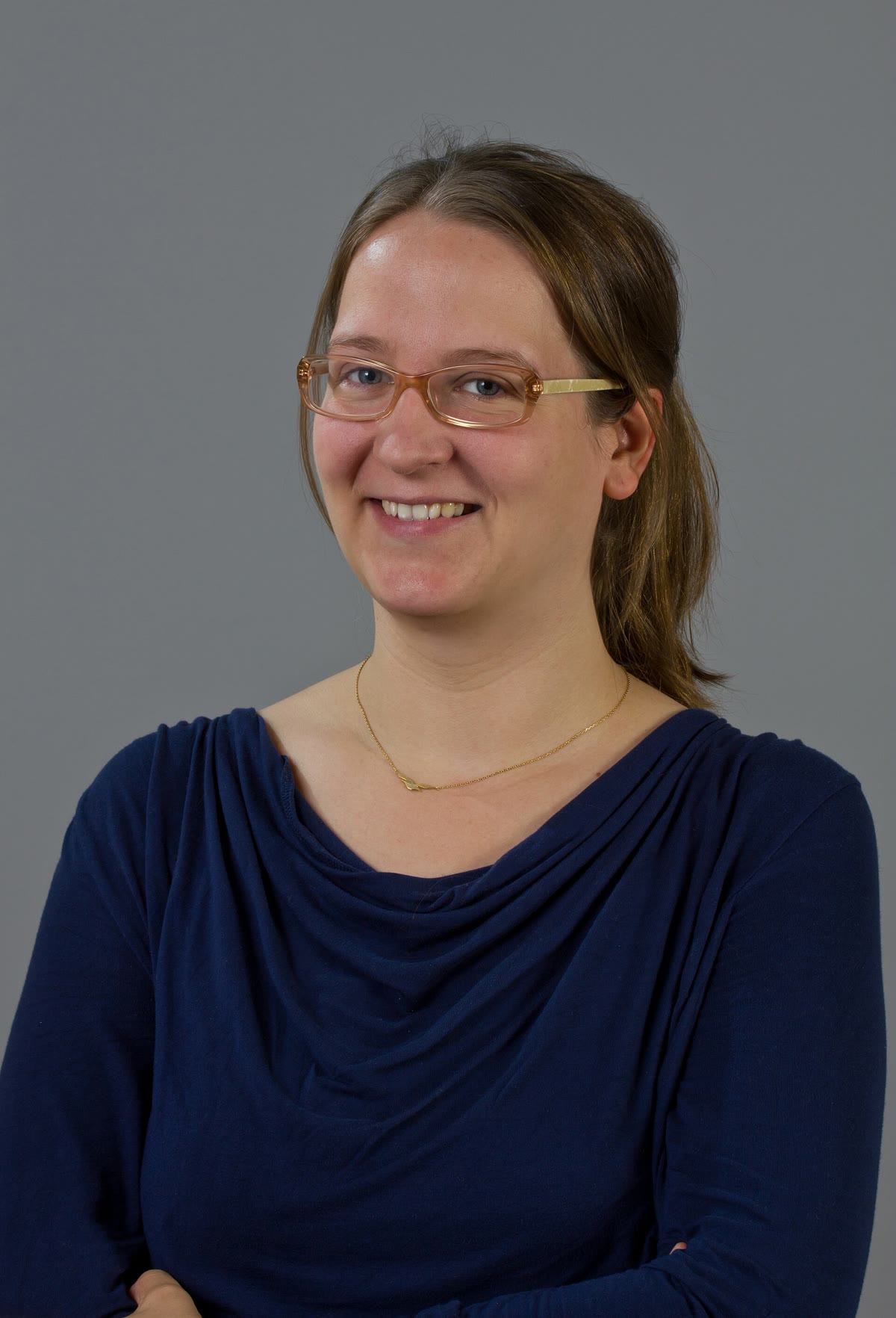 A studio portrait of a woman with glasses wearing a deep navy draped cowl-neck top and a delicate gold necklace