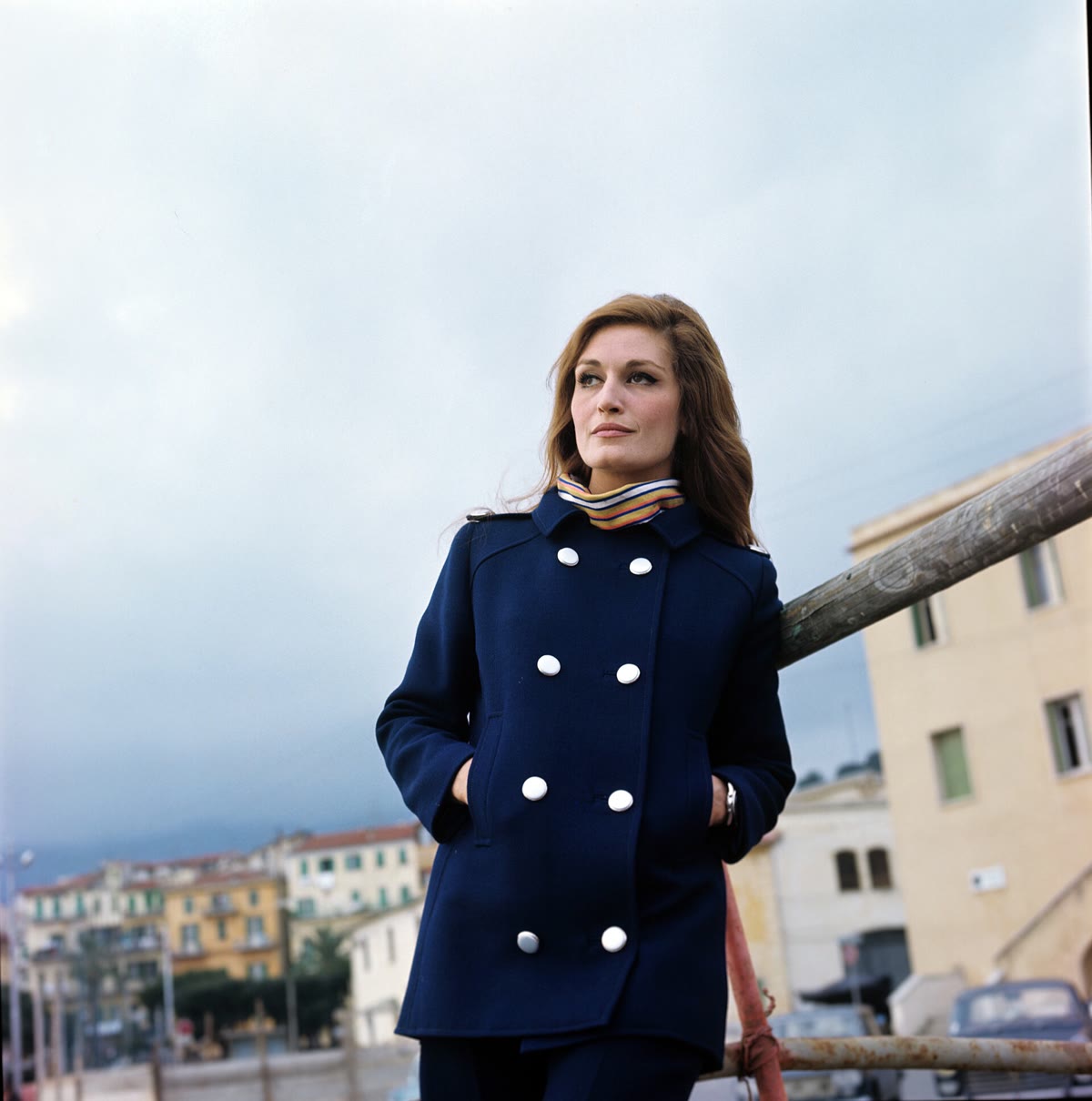A woman leaning on a railing outdoors wearing a deep navy double-breasted peacoat with large white buttons over a striped turtleneck