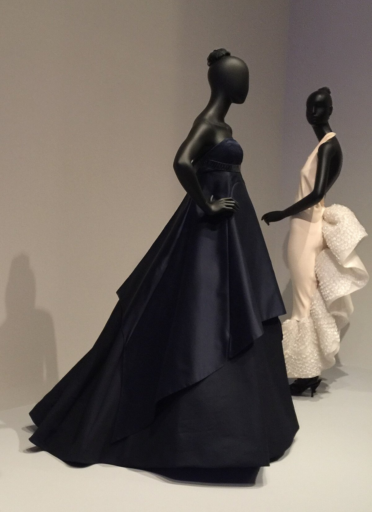 A deep navy strapless satin evening ball gown with a full sweeping skirt displayed on a black museum mannequin next to a white gown