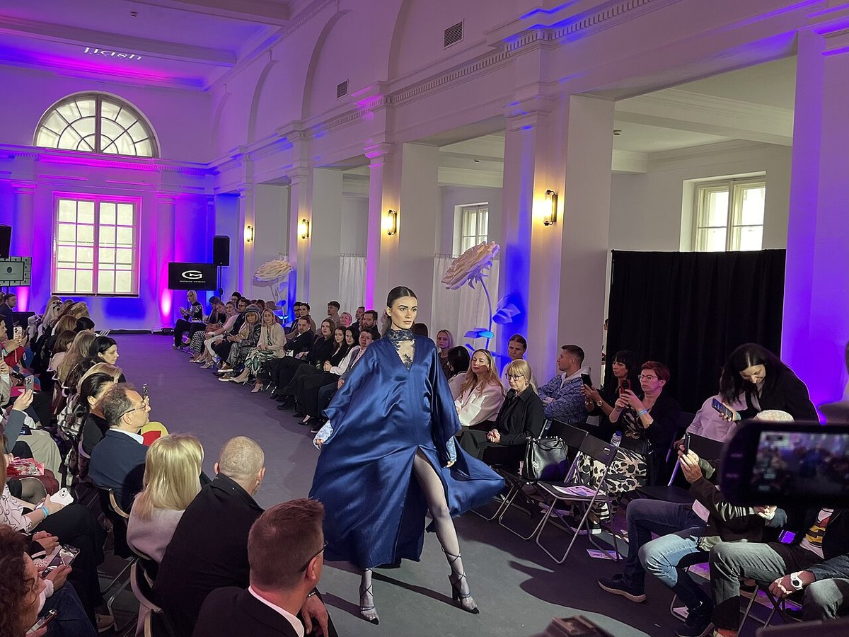 A model walking a fashion runway in a flowing deep navy satin gown with a thigh-high slit, in front of a seated audience under purple-blue lighting