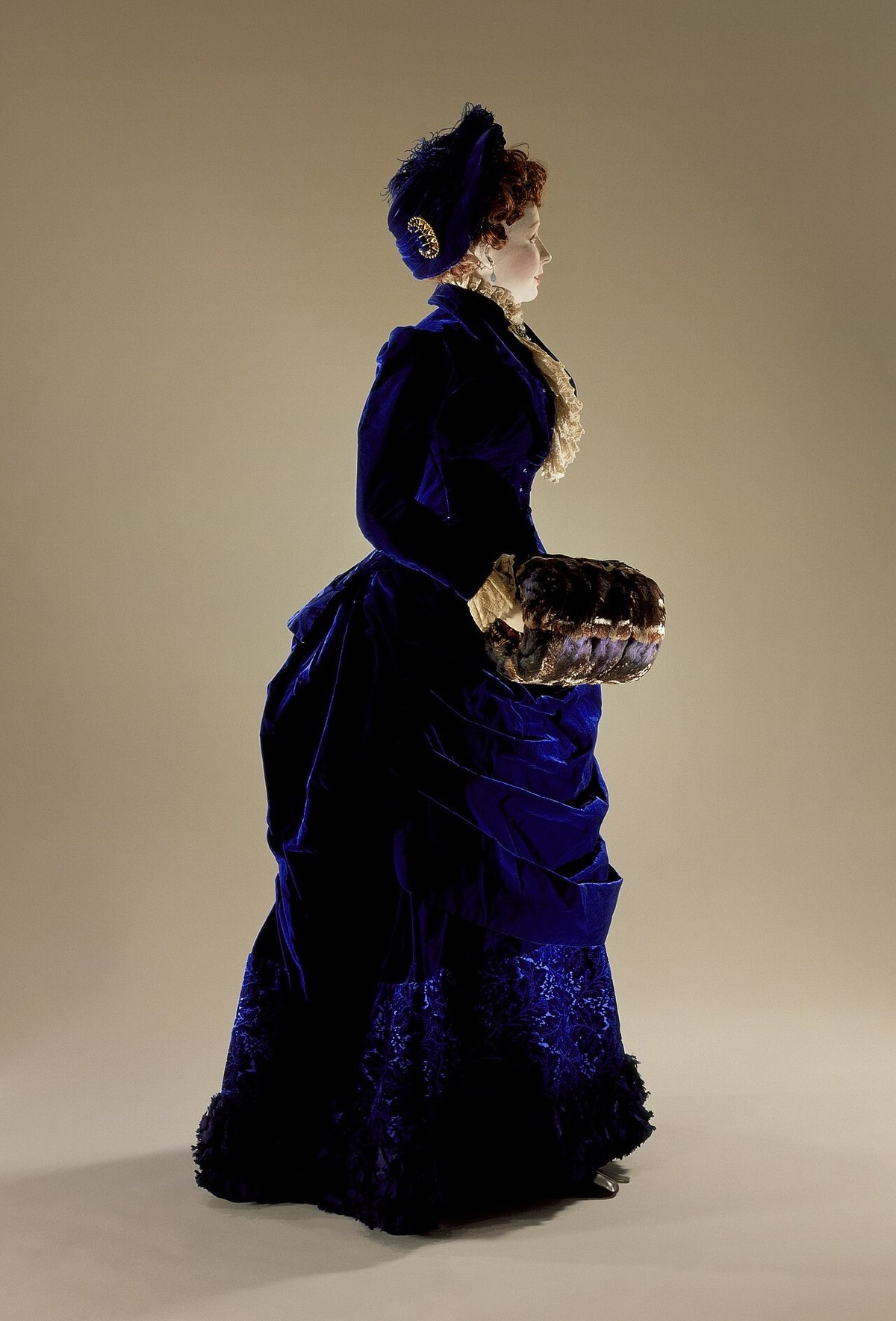 An 1880s deep blue silk velvet dress ensemble with bustle, lace jabot, matching hat, and fur muff displayed on a mannequin