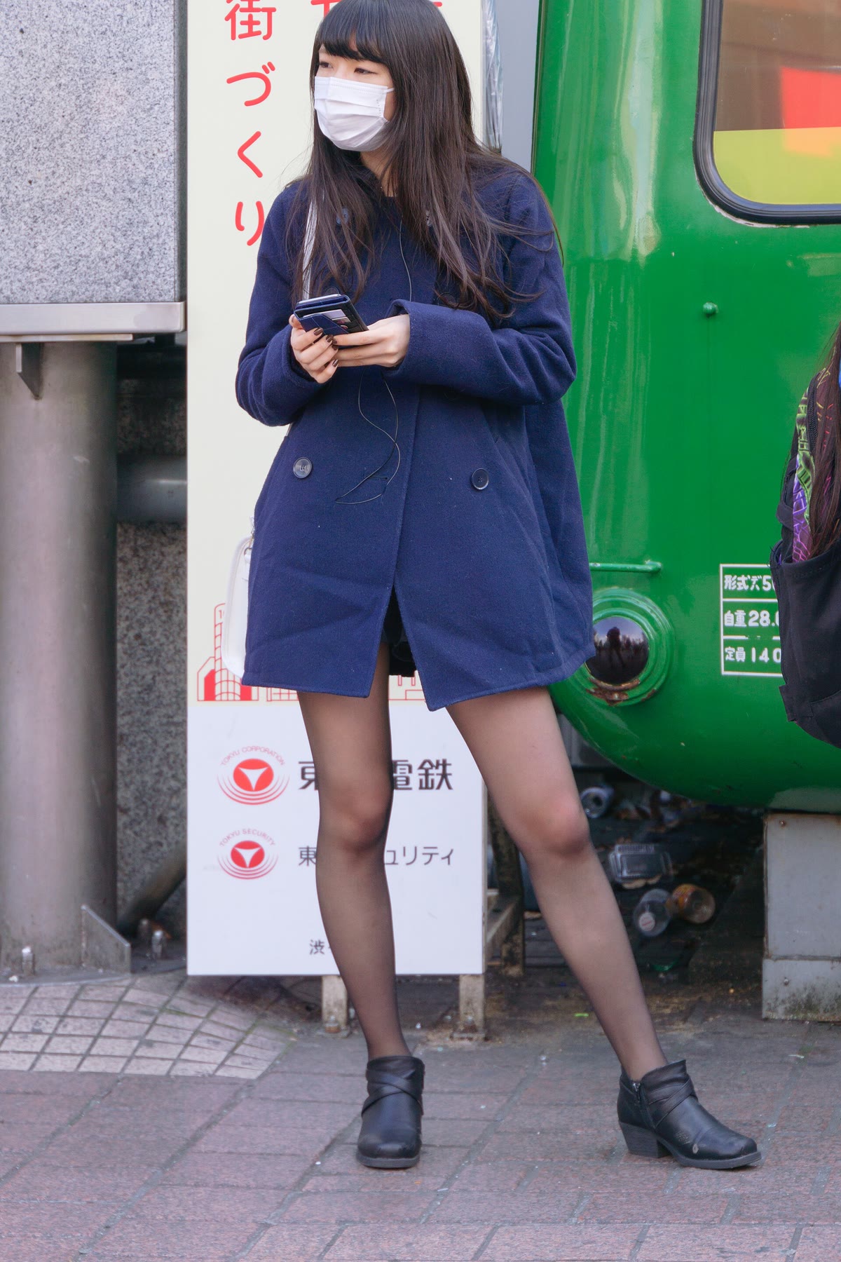 A young woman standing at a city station wearing a deep navy wool coat, black tights, and black ankle boots, looking at her phone