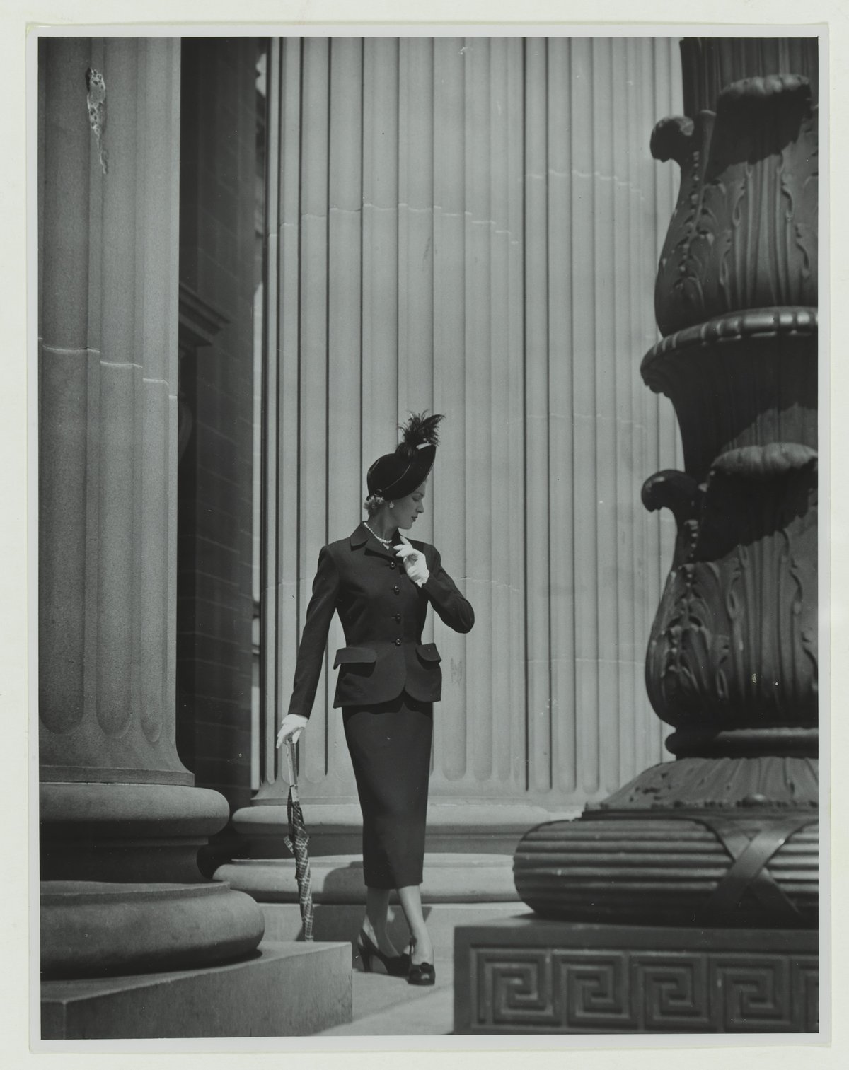 Vintage black-and-white photograph of a model in a dark New Look skirt suit and feathered hat descending stone steps
