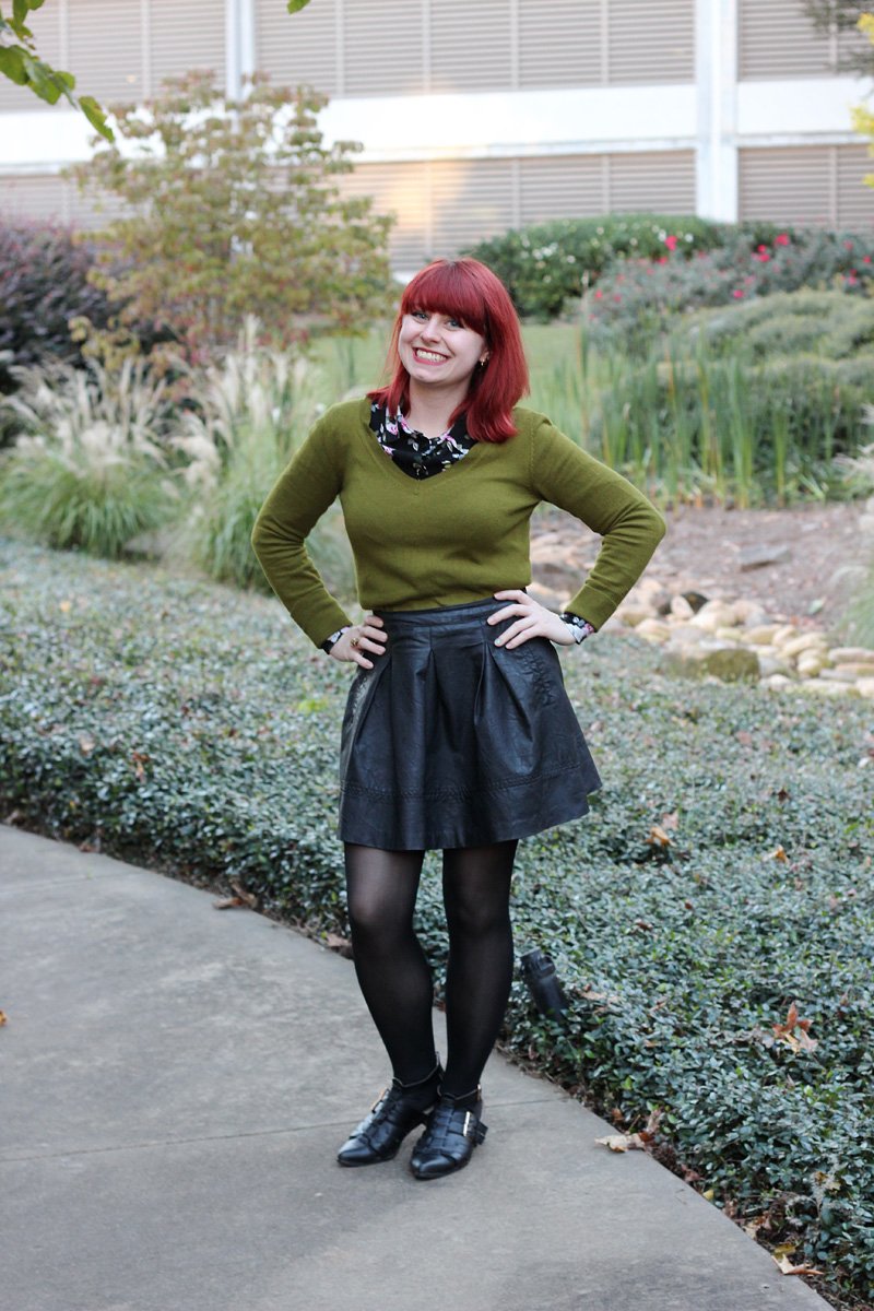 A woman with red hair wearing an olive green V-neck sweater over a black floral shirt, a black faux-leather skater skirt, black tights and cutout boots