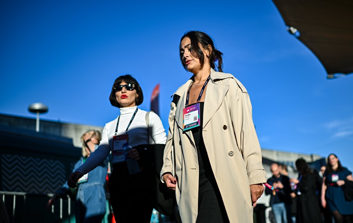 A woman at an outdoor event wearing an open beige trench coat over a black dress, with an attendee lanyard around her neck