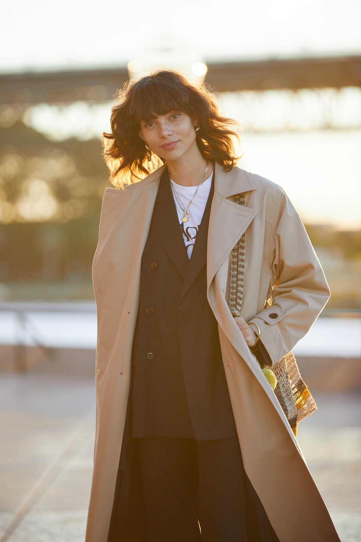 A model at golden hour wearing an open beige trench coat over a dark double-breasted blazer, white graphic tee and layered necklaces, carrying a woven bag