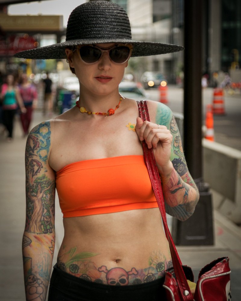A tattooed woman on a Minneapolis street wearing a bright orange bandeau top, black wide-brim hat and sunglasses, black bottoms, a red bag and an orange beaded necklace