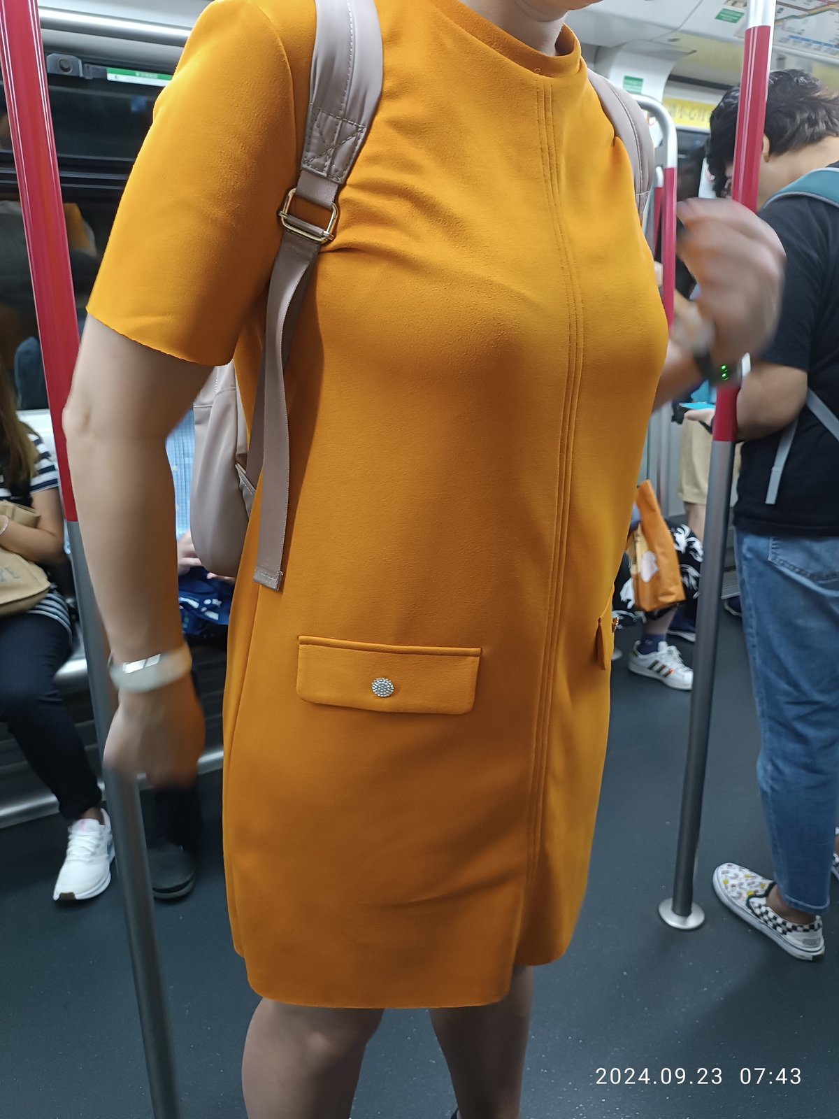 A woman standing in a Hong Kong MTR train wearing a pumpkin-orange suede short-sleeve mini shift dress with a front seam, patch pockets with crystal buttons, and a beige backpack