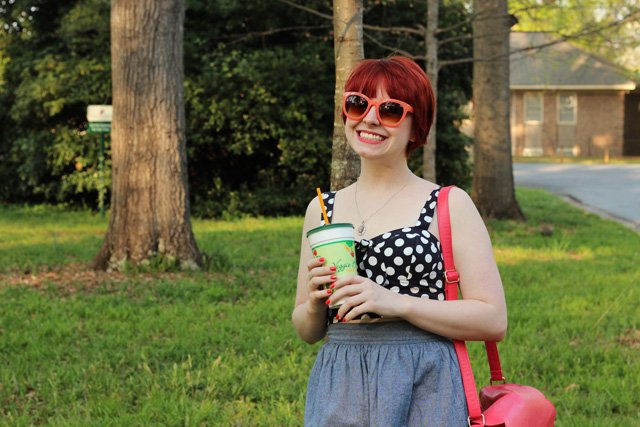 A woman with a red pixie cut wearing orange sunglasses, a black-and-white polka dot top, a blue skirt and a coral crossbody bag, holding a drink outdoors