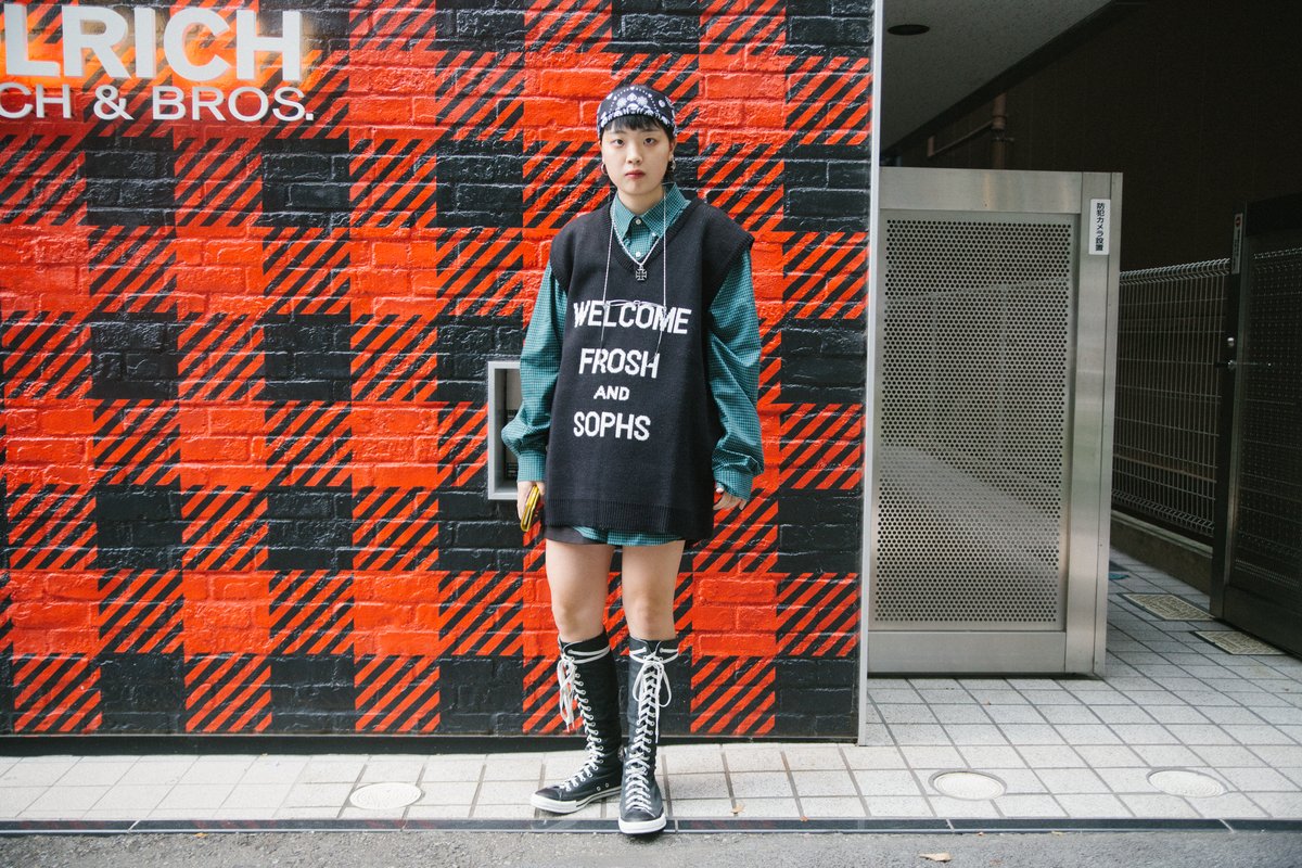 Person in an oversized red-and-black plaid coat and boots standing by a plaid wall in Tokyo