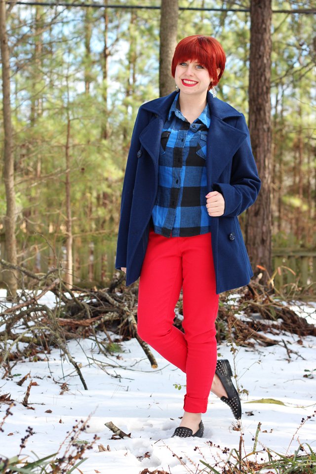 A woman with a red pixie haircut wearing a blue peacoat over a plaid shirt, red jeans and studded loafers, standing in a snowy wooded area