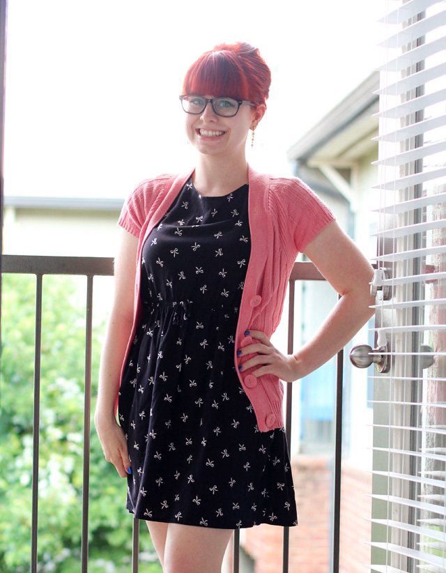 A woman with red hair and glasses wearing a pink cable-knit short-sleeve cardigan over a black dress with a white bow print, standing on a balcony